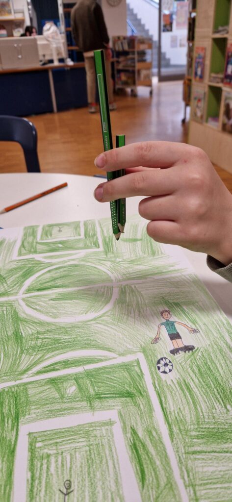 A hand holds two green colored pencils above a drawing of a soccer field with a cartoon boy and a soccer ball. The table and shelves with books are visible in the background.