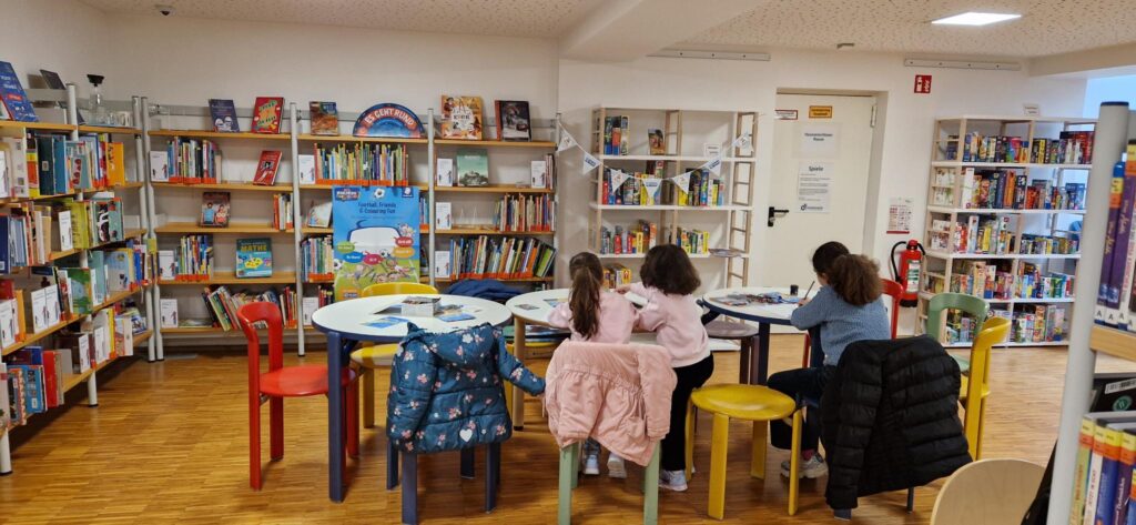 Children sit at round tables in a colorful library, reading and drawing. Shelves filled with books and board games line the walls. Coats hang on the backs of the chairs, and the room appears bright and inviting.