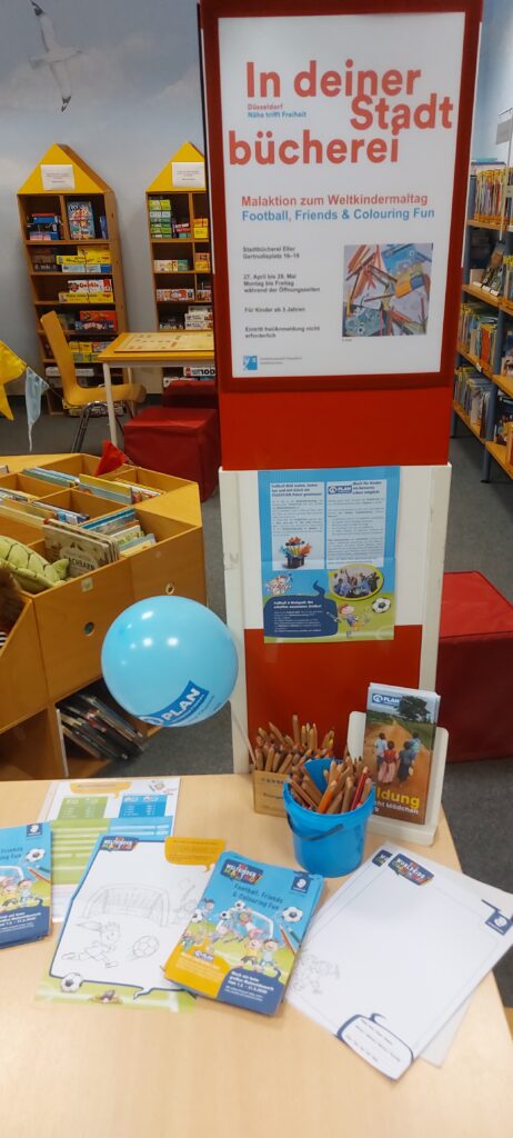 A library display features a sign promoting a football and coloring event for World Childrens Day. On a table are coloring sheets, crayons, flyers, a blue balloon, and a cup of pencils. Bookshelves are visible in the background.