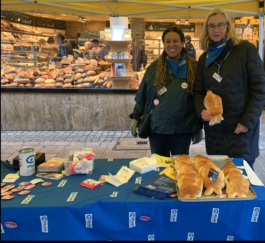 Vor einer Bäckerei stehen zwei Frauen hinter einem mit einem blauen Tuch bedeckten Tisch und bieten Backwaren und Informationsmaterial an.