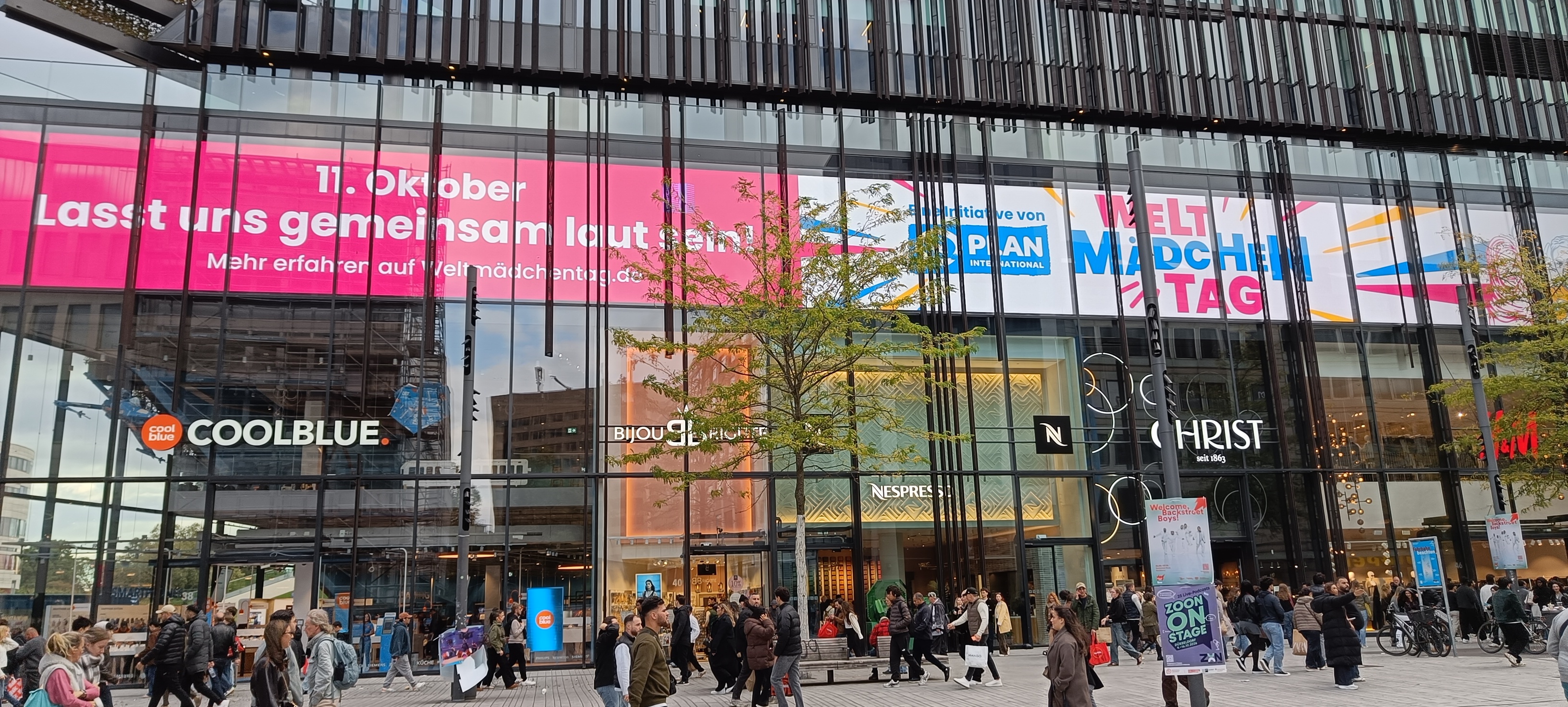 Außenansicht eines modernen Geschäftsgebäudes mit großen Glasfenstern, Ladenfronten von Coolblue, Nespresso und Christ sowie einem großen rosa Banner, das für den Internationalen Mädchentag am 11. Oktober wirbt.