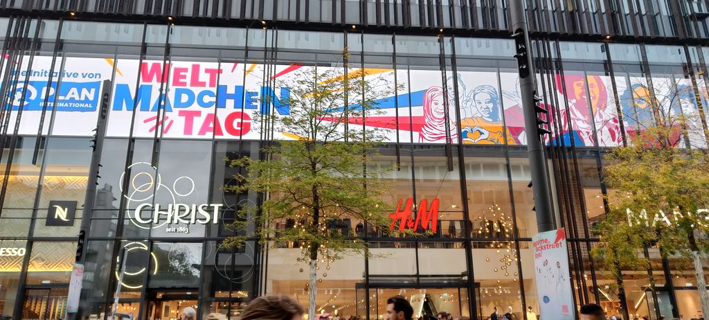 Eine große Glasfassade mit einem Banner, auf dem Welt-Mädchen-Tag steht, sowie Illustrationen von Mädchen und Logos von Plan International, Christ und H&M.
