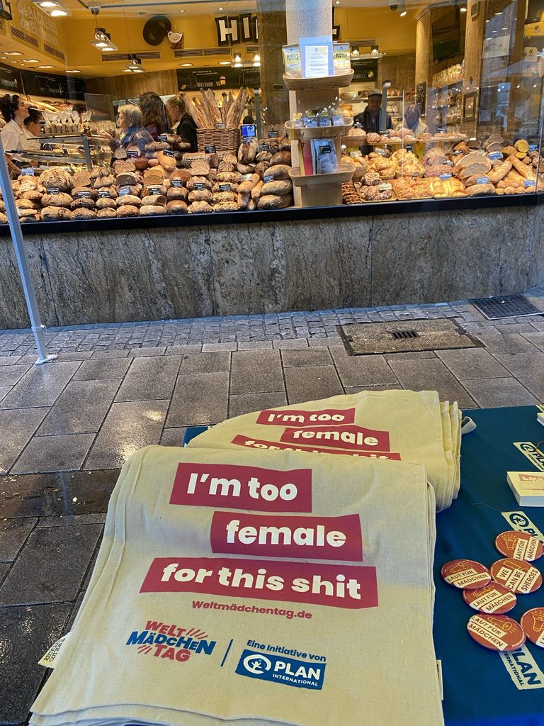 Aufgestapelte gelbe Tragetaschen mit den Aufschriften Ich bin zu weiblich für diesen Mist und Weltmädchentag vor dem Schaufenster einer Bäckerei.