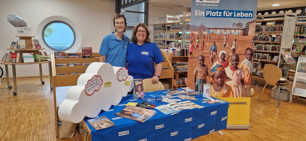 Zwei Personen in identischen blauen Hemden stehen hinter einem mit einem blauen Tuch bedeckten Tisch, im Hintergrund ist ein großes Plan-International-Banner mit Fotos von Kindern zu sehen.