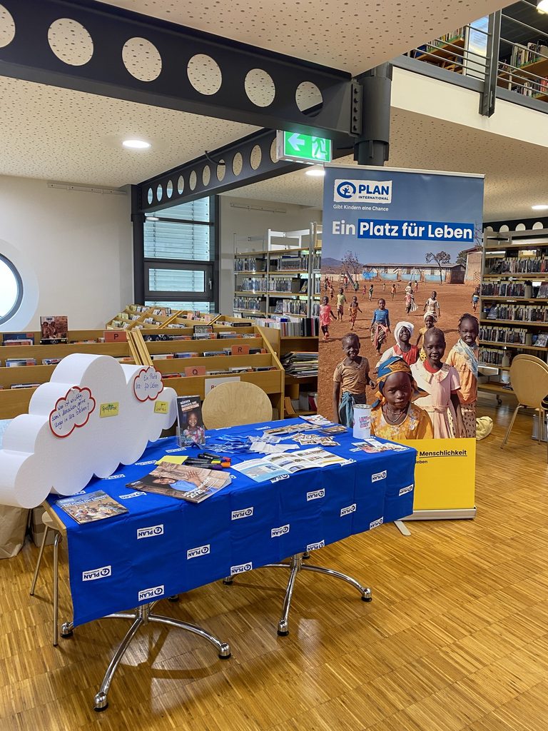 Ein Informationsstand von Plan International in einer Bibliothek mit Broschüren und einem hohen Banner, auf dem Fotos von Kindern in Afrika sowie der Text Ein Platz für Leben zu sehen sind.