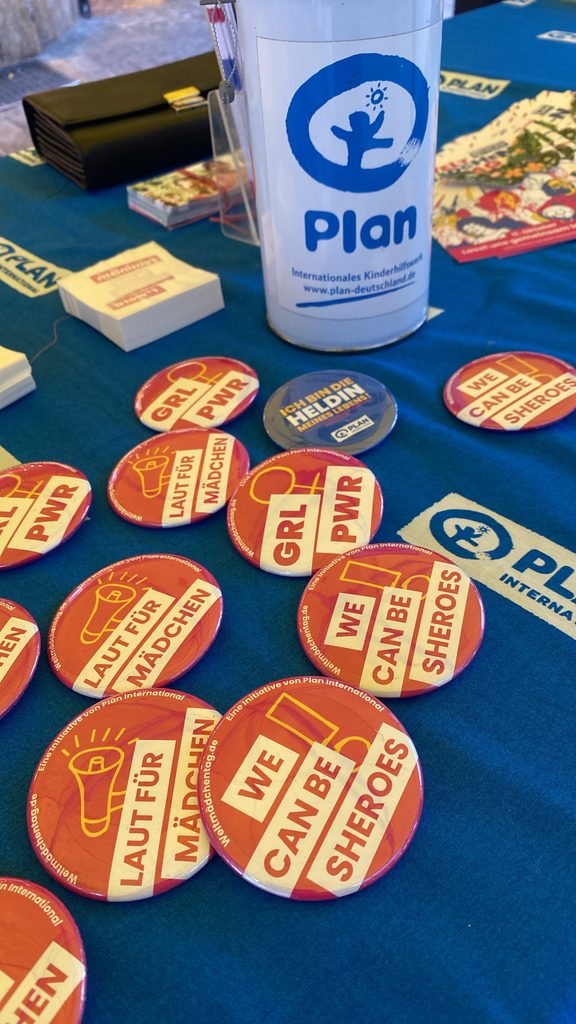 Eine Sammlung bunter Ansteckbuttons mit Slogans wie GRL PWR, WE CAN BE SHEROES und LAUT FÜR MÄDCHEN auf einem blauen Tisch neben einer Sammelbüchse von Plan International.