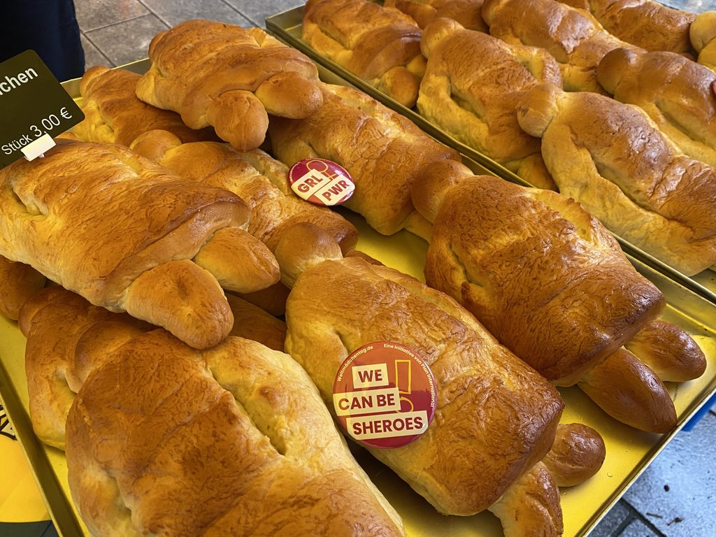 Ein Tablett mit goldbraunen, schildkrötenförmigen Brötchen, von denen einige mit Aufklebern mit den Aufschriften GRL PWR und WE CAN BE SHEROES verziert sind; daran hängt ein Preisschild mit der Aufschrift 3,00 €.
