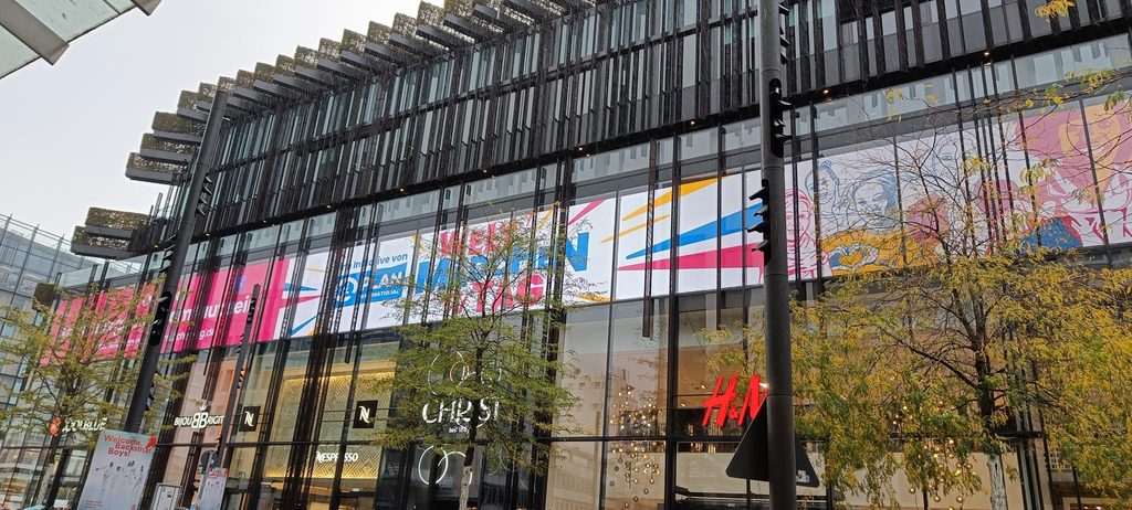 Modernes Einkaufszentrum mit Glasfassade, in dem sich H&M- und Nespresso-Filialen befinden, sowie einem großen, farbenfrohen Werbebanner für Plan International.